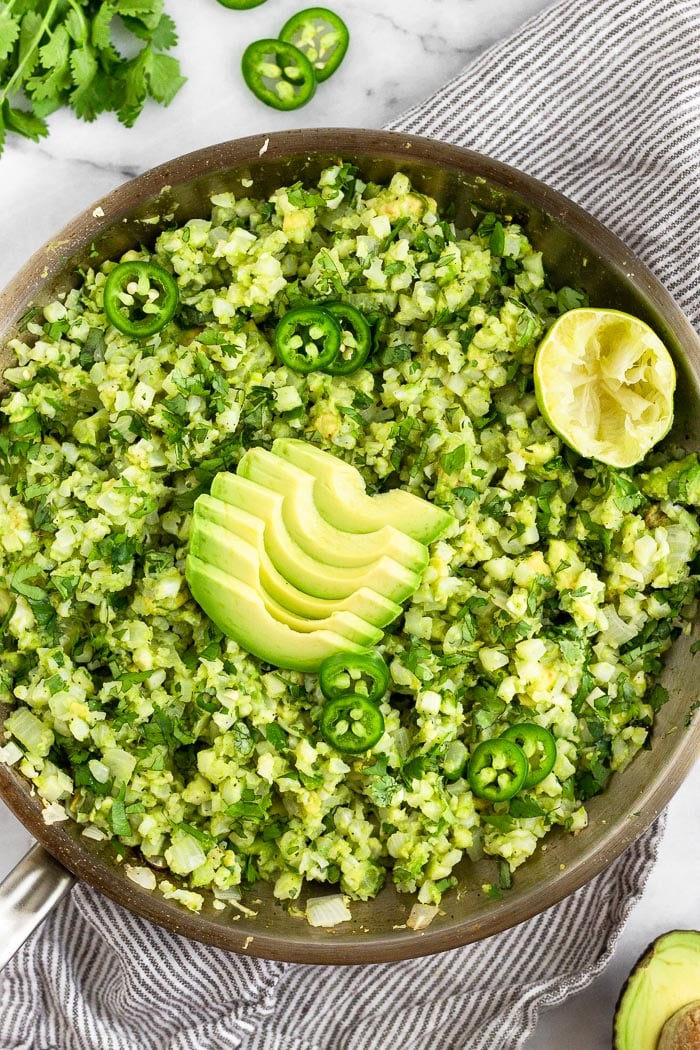 A large pan filled with avocado cauliflower rice topped with sliced jalapeños, avocado slices, and half a lime. The pan is sitting on a towel and around it is half and avocado, cilantro, and sliced japaleons.