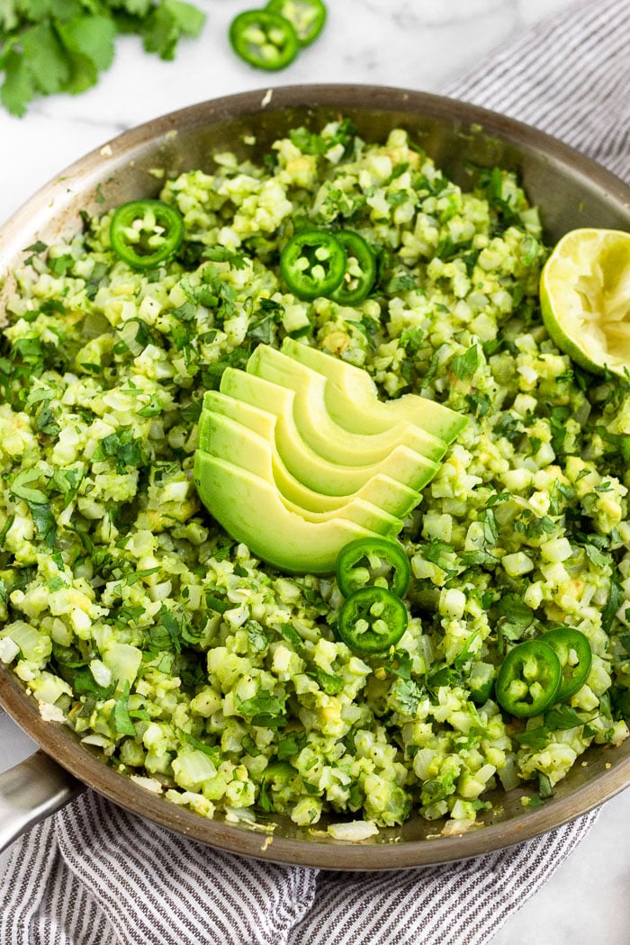 Large pan filled with avocado cauliflower rice topped with sliced avocado, sliced jalapenos, cilantro, and half a juiced lime. Behind it is more cilantro and sliced jalapeños.