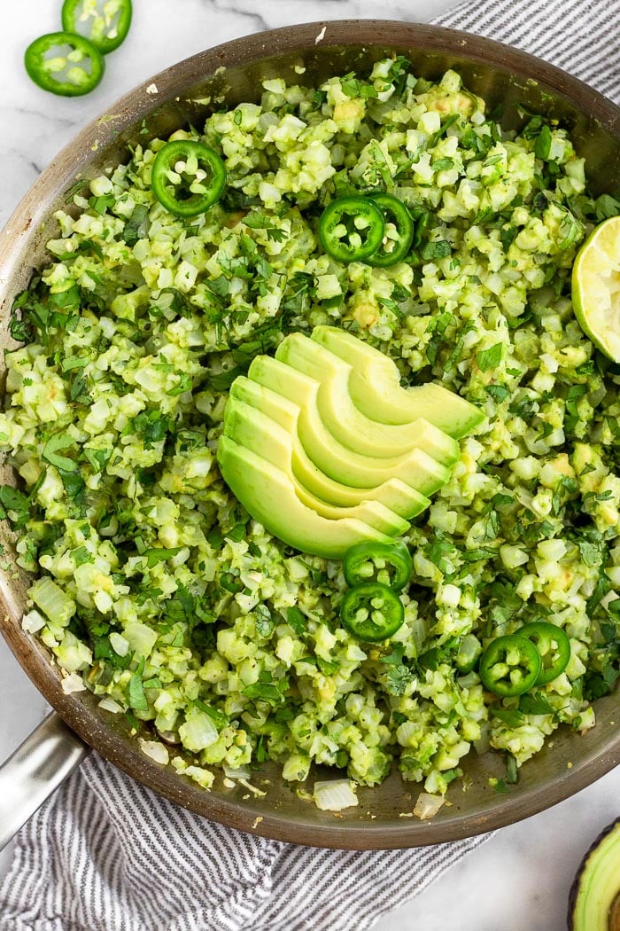 Large pan of avocado cauliflower rice. It is garnished with avocado slices. sliced jalapeños, and half a juice lime. Around it is a stripped towel, half and avocado, and sliced jalapeños.