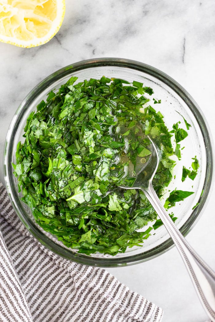 Bowl of fresh parley, cilantro, lemon juice, and olive oil all mixed up with a spoon coming out of it.