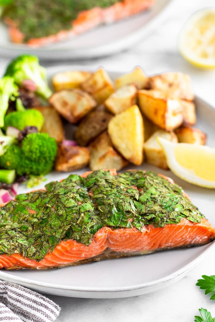 Herb grilled salmon on a plate with potatoes and broccoli and a lemon wedge for garnish. Behind it is another plate of salmon and half a lemon.