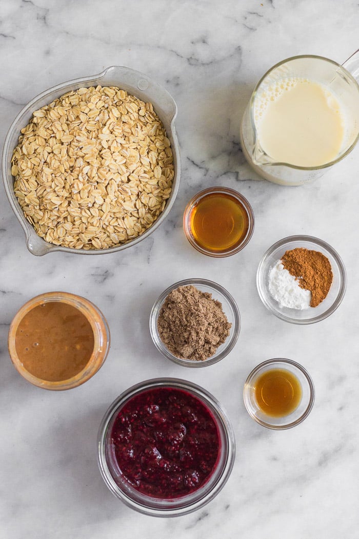 Overhead shot of different small bowls filled with rolled oats, almond milk, maple syrup, spices, ground flax, vanilla extract, peanut butter, and jelly.