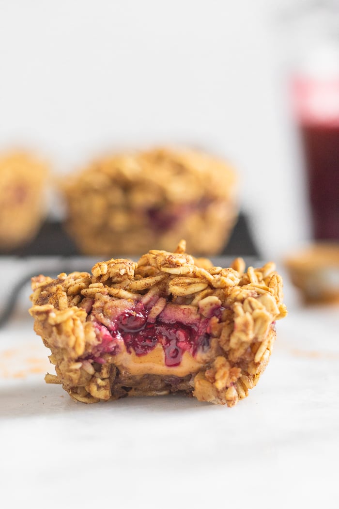 Baked oatmeal cup with a bite taken out of it with peanut butter and jelly coming out. Behind it is a cooling rank with more muffins, a jar of jam, and a spoon of peanut butter.
