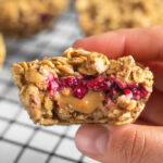 A hand holding a peanut butter and jelly stuffed oatmeal cup with a bite taken out of it. Behind it is more on a baking rake.
