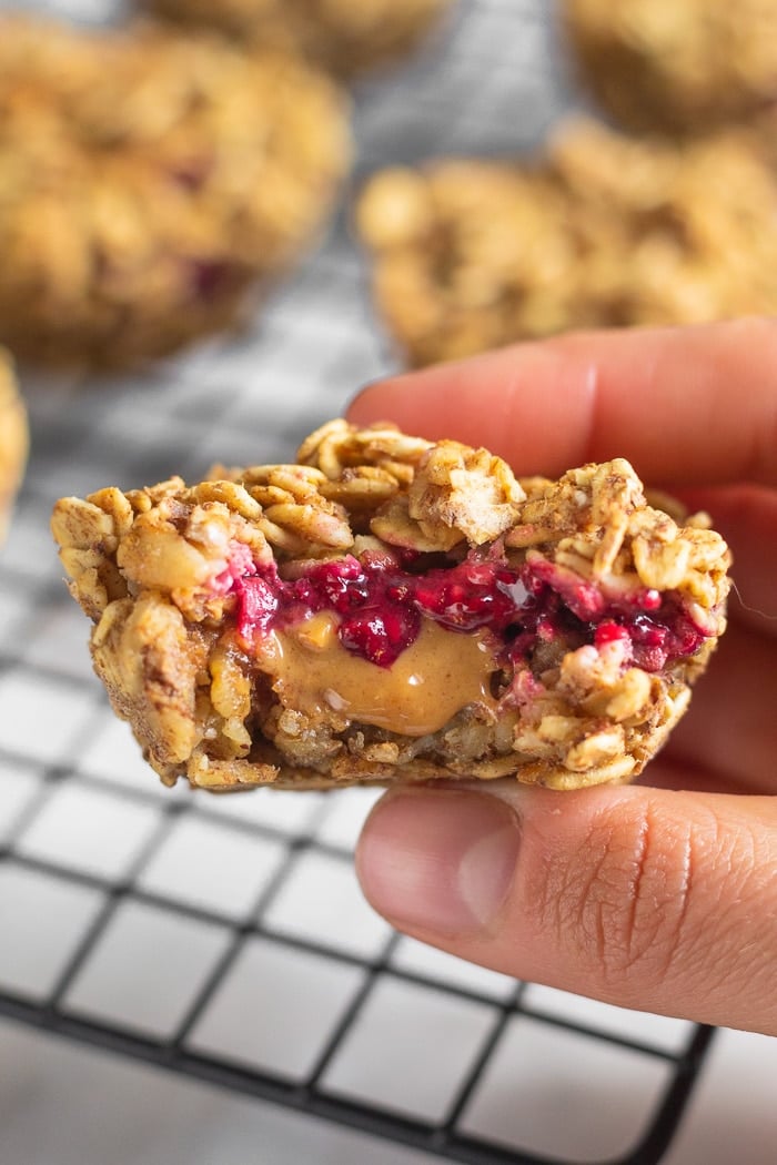 A hand holding a peanut butter and jelly stuffed oatmeal cup with a bite taken out of it. Behind it is more on a baking rake.