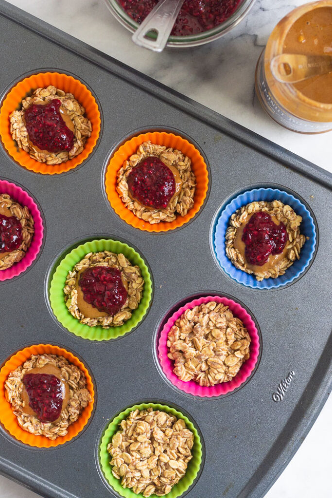 Silicone muffin liners filled with rolled oats and then some are topped with peanut and then jelly before more rolled oat mixture is placed on top. Next to it a jar of jam and a jar of peanut butter.