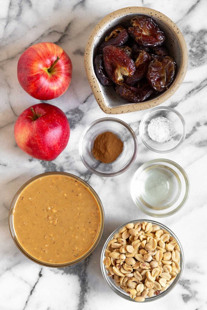 While marble counter with a bowl of medjool dates, a bowl of sea salt, a bowl of cinnamon, a bowl of coconut oil, a bowl of peanuts, a bowl of peanut butter, and two red apples.