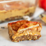 Close up of an apple peanut butter bar sitting on a white counter. Behind it is a pan of more bars, a spoon with peanut butter on it, and an apple.