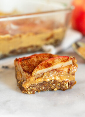 Close up of an apple peanut butter bar sitting on a white counter. Behind it is a pan of more bars, a spoon with peanut butter on it, and an apple.