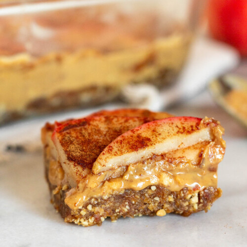 Close up of an apple peanut butter bar sitting on a white counter. Behind it is a pan of more bars, a spoon with peanut butter on it, and an apple.