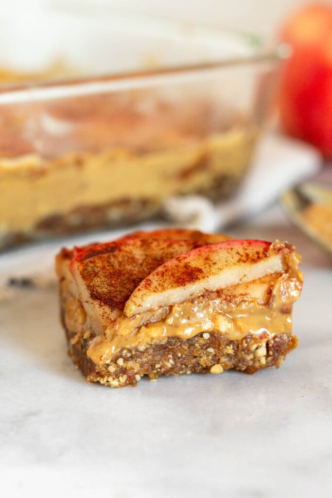 Close up of an apple peanut butter bar sitting on a white counter. Behind it is a pan of more bars, a spoon with peanut butter on it, and an apple.