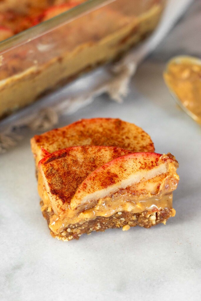 3 layer peanut butter apple bar on a white counter top. Behind the bar is a pan with more bars and a spoon with peanut butter on it.