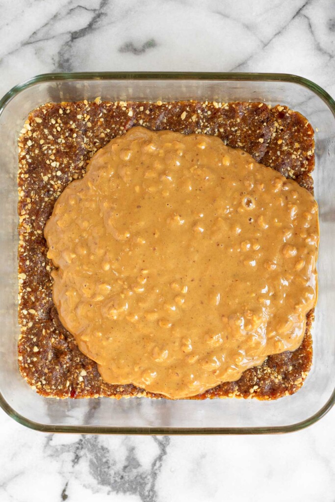 A baking dish with a sticky date and nut mixture pressed into the bottom of the pan. A layer of crunchy peanut butter is on top of it before it is spread out.