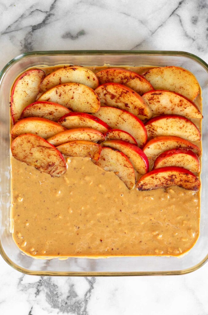 Sautéed sliced apples being layered on top of crunchy peanut butter in a glass baking dish.
