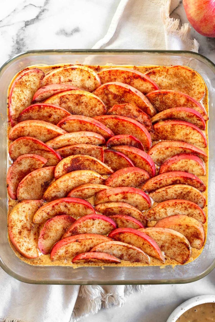 An overhead shot of a baking dish filled with apple bars. The dish is sitting on a white towel with an apple and a bowl of peanut butter next to the dish.