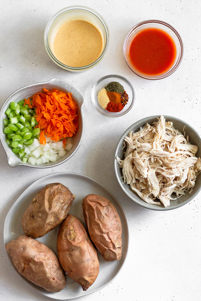 Overhead shot of a jar of mayo, bowl of hot sauce, bowl os shredded chicken, plate of cooked sweet potatoes, bowls of veggie mixture, and a bowl of spices.