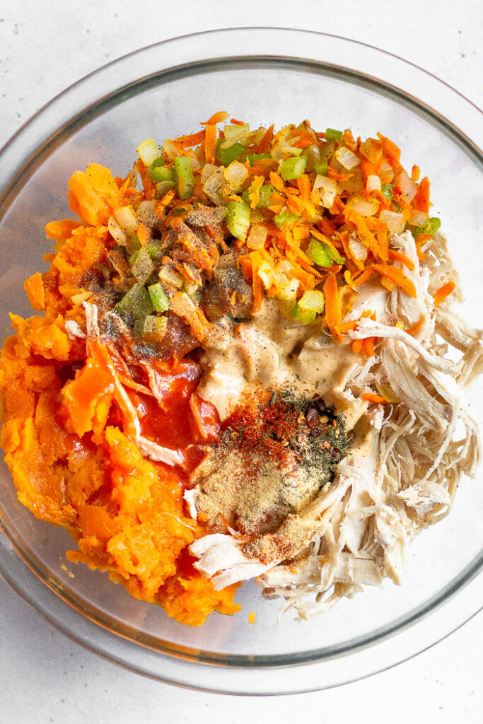 Large glass bowl filled with shredded chicken, mashed sweet potato, veggies, mayo, hot sauce, and spices.