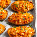 Buffalo chicken stuffed sweet potatoes on a baking sheet drizzled with sauce and topped with fresh herbs.