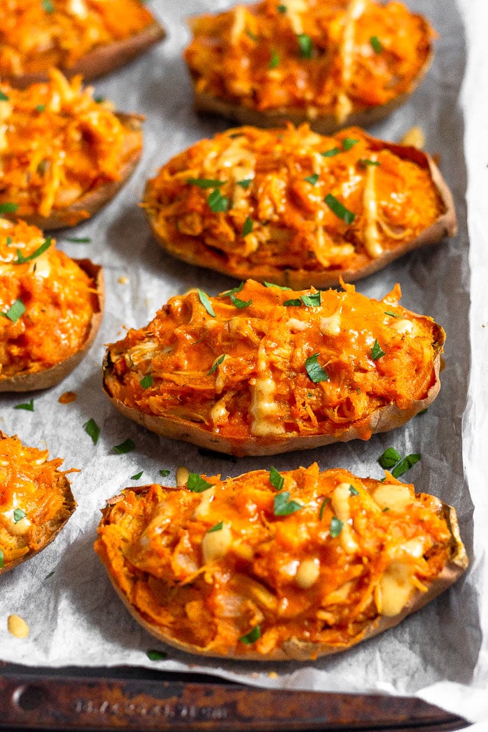 Buffalo chicken stuffed sweet potatoes on a baking sheet drizzled with sauce and topped with fresh herbs.