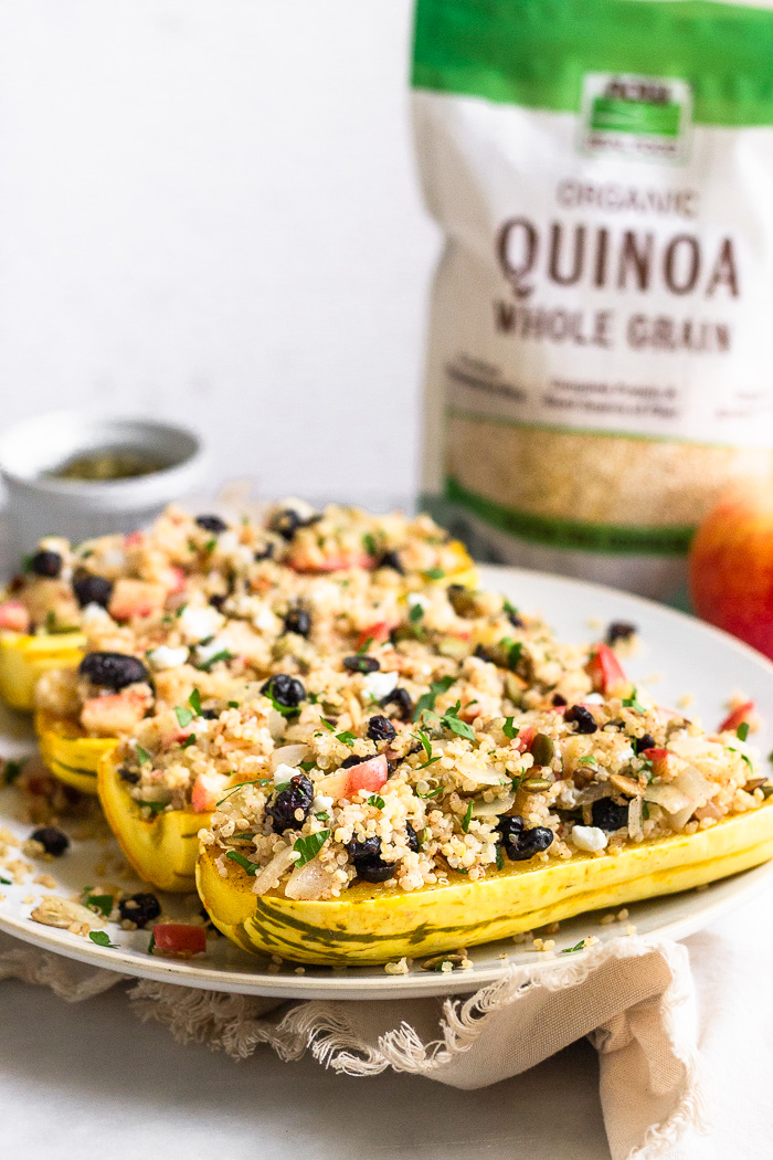 Quinoa stuffed squash on a plate. Behind it is an apple, a bag of quinoa, and a small bowl of pumpkin seeds.