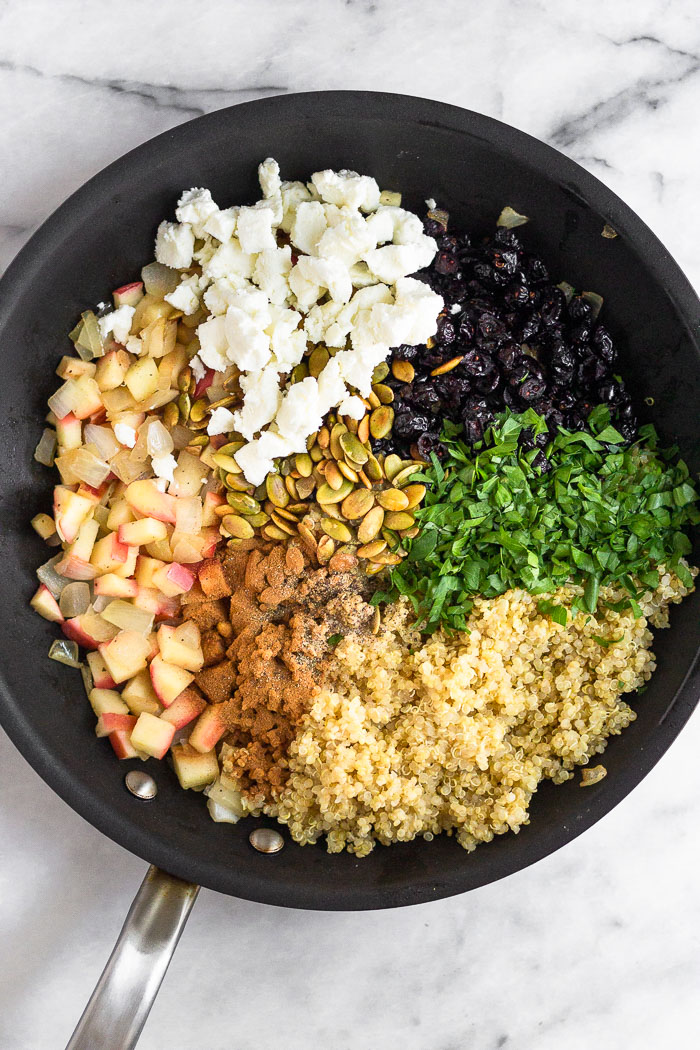 Saute pan filled with different piles of sautéed apples and onions, cooked quinoa, chopped parsley, cranberries, goat cheese, pumpkin seeds, and spices.