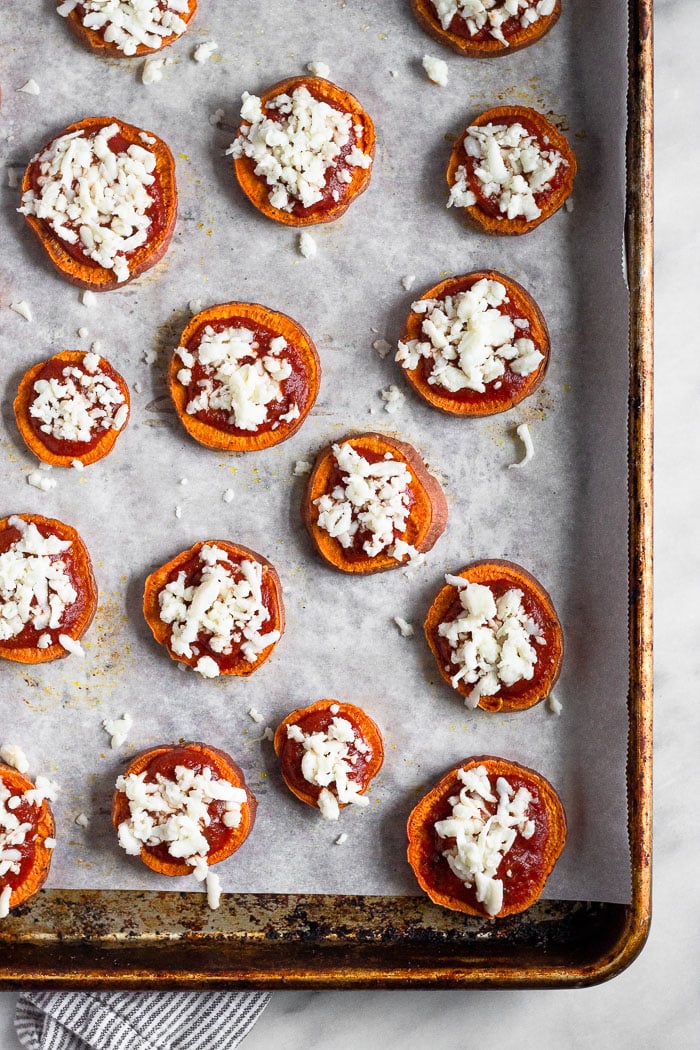A baking sweet lined with parchment paper with cooked sweet potato rounds topped with sauce and shredded cheese before being placed back in the oven.