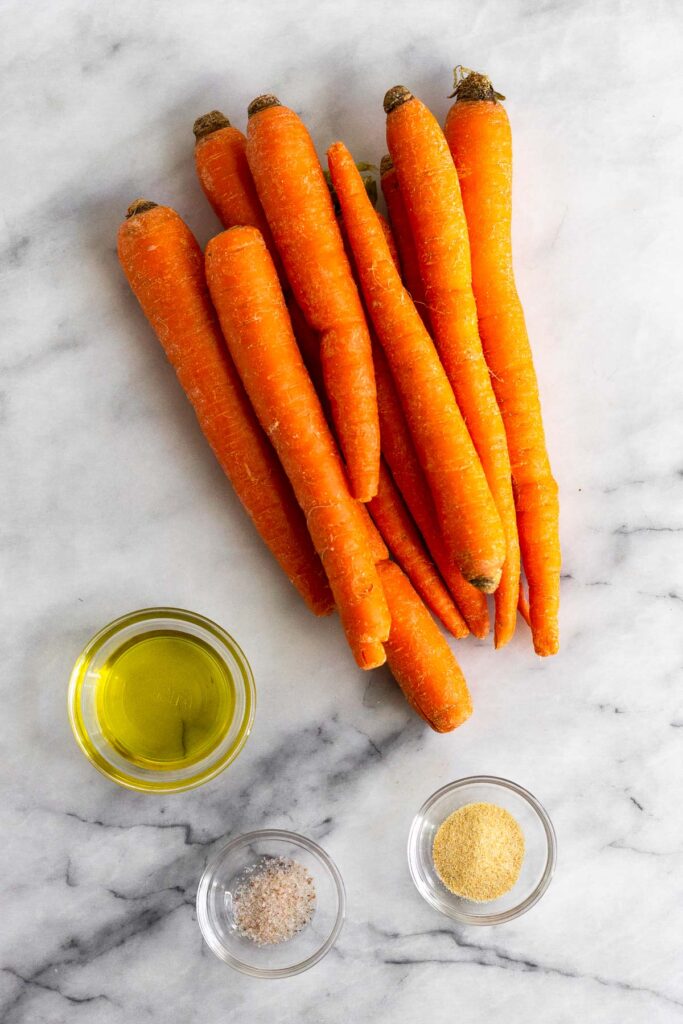 Marble counter with a pile of large carrots, a bowl of garlic powder, a bowl of salt, and a bowl of olive oil.