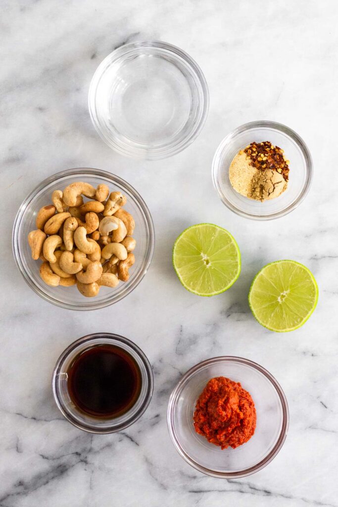 Marble counter with a bowl of water, a bowl of spices, 2 halves of limes, a bowl of red curry paste, a bowl of coconut aminos, and a bowl of cashews.