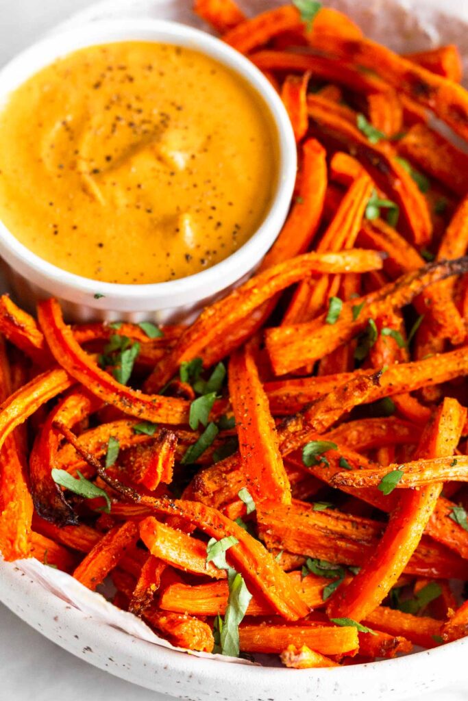 Close up of carrot fries topped with cilantro and a small ramekin of orange sauce all in a white bowl.