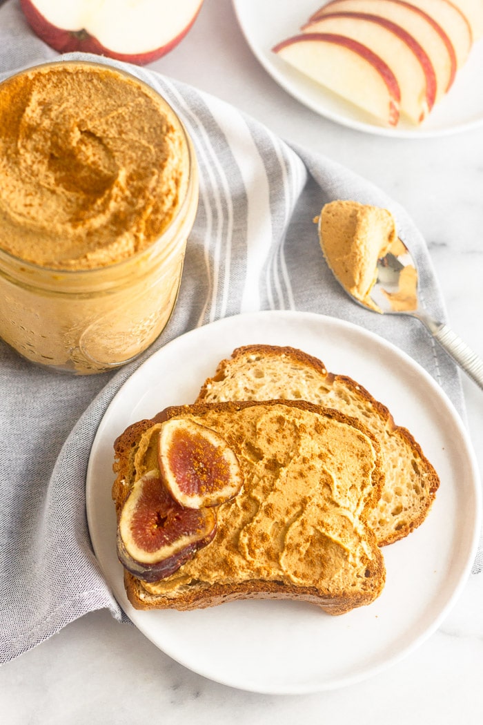Two pieces of toast on a plate with pumpkin butter spread on one and sliced figs. Next to it is a jar of more pumpkin butter with a spoon and apple slices in the background.