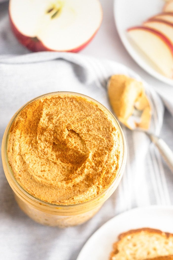 Pumpkin pie butter in a jar surrounded by half an apple, a plate of apple slices, and a spoon with more pumpkin butter on it.