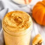 Jar filled with homemade pumpkin pie butter sprinkled with cinnamon. Next to it is a spoon with some on it, a pumpkin, and a plate of sliced apples in the back.