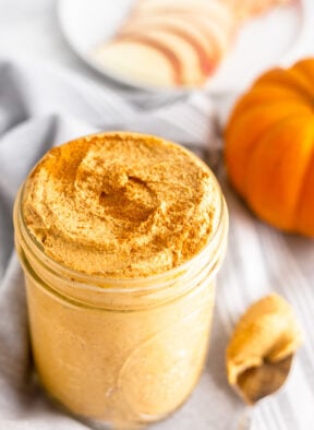 Jar filled with homemade pumpkin pie butter sprinkled with cinnamon. Next to it is a spoon with some on it, a pumpkin, and a plate of sliced apples in the back.