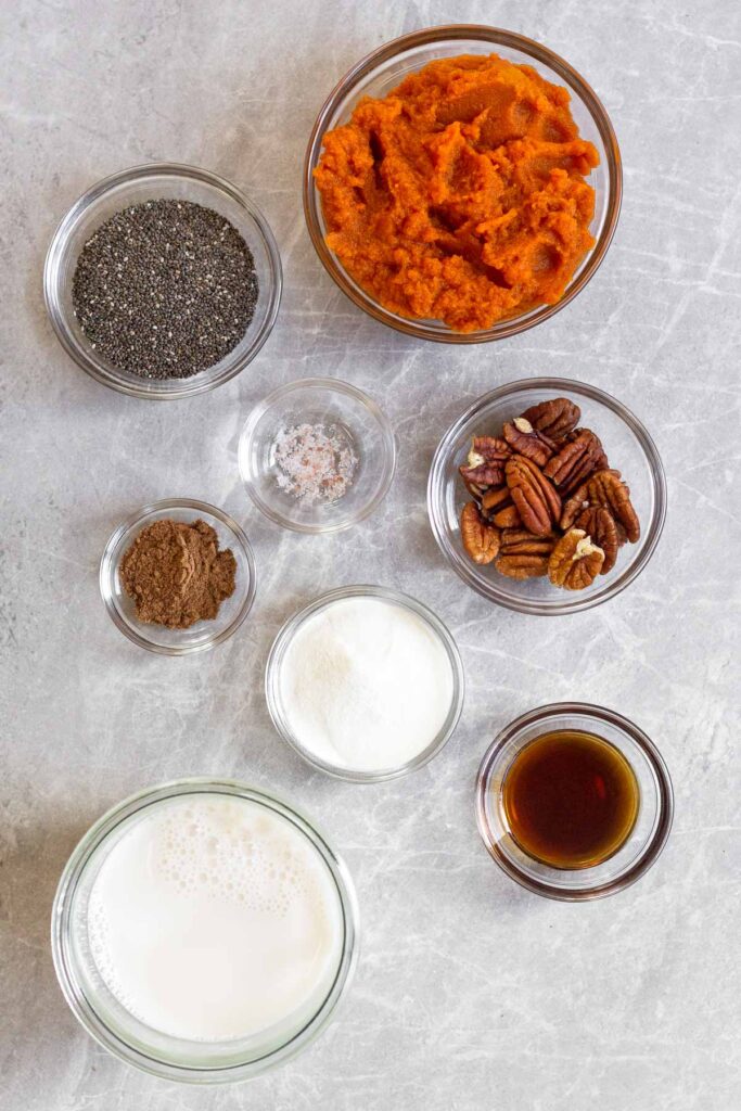 Overhead shot of a bowl of pumpkin puree, bowl of pecans bowl of maple syrup, jar of milk, bowl of collagen, bowl of spices, bowl of salt, and a bowl of chia seeds.