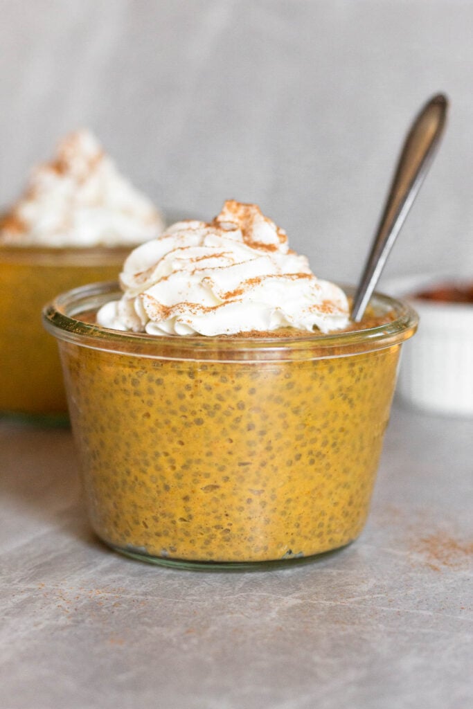 A jar filled with pumpkin pie chia pudding topped with whipped cream and cinnamon with a spoon in it. Behind it is another jar.