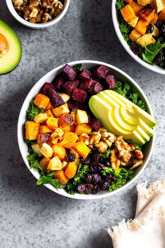 Butternut squash power bowl with kale, beets, parsnips, avocado, walnuts, and dried cranberries, all topped with a tahini dressing. Next to it is a kitchen towel, another bowl, container of walnuts, and half an avocado.