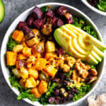 Veggie power bowl with kale, butternut squash, parsnips, beets, avocado, walnuts, and dried cranberries. It is topped with tahini dressing. Next to is is another bowl, half an avocado, bowl of walnuts, and a kitchen towel.