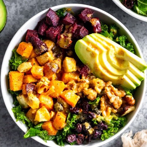 Veggie power bowl with kale, butternut squash, parsnips, beets, avocado, walnuts, and dried cranberries. It is topped with tahini dressing. Next to is is another bowl, half an avocado, bowl of walnuts, and a kitchen towel.