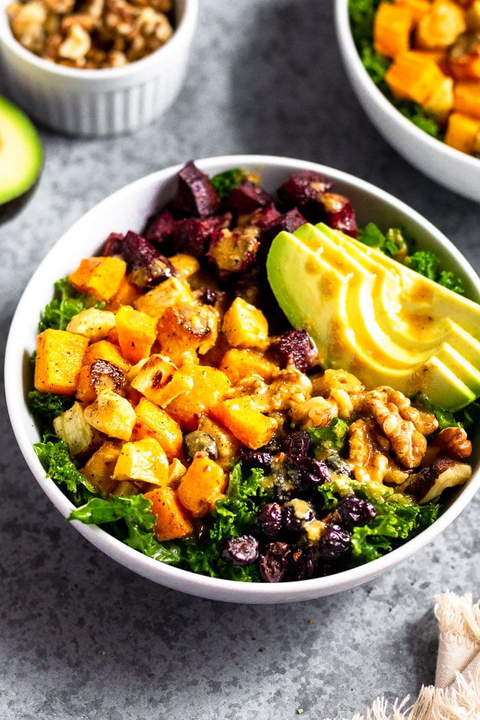 Winter power salad with roasted squash, parsnips, and beets, avocado, nuts, and dried fruit. It is topped with a vegan tahini dressing. Behind it is another bowl, dish of walnuts, and half an avocado.