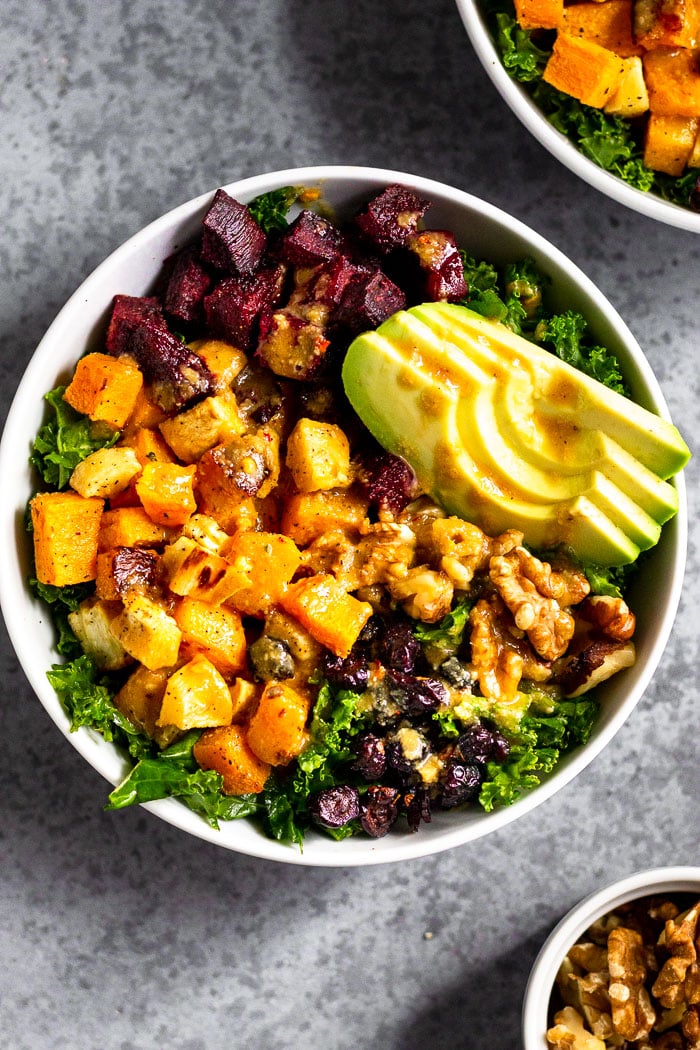 Vegan power bowl filled with butternut squash, parsnips, beets, kale, avocado, walnuts, and dried cranberries and topped with a tahini dressing. Next to is is another bowl and small dish of walnuts.