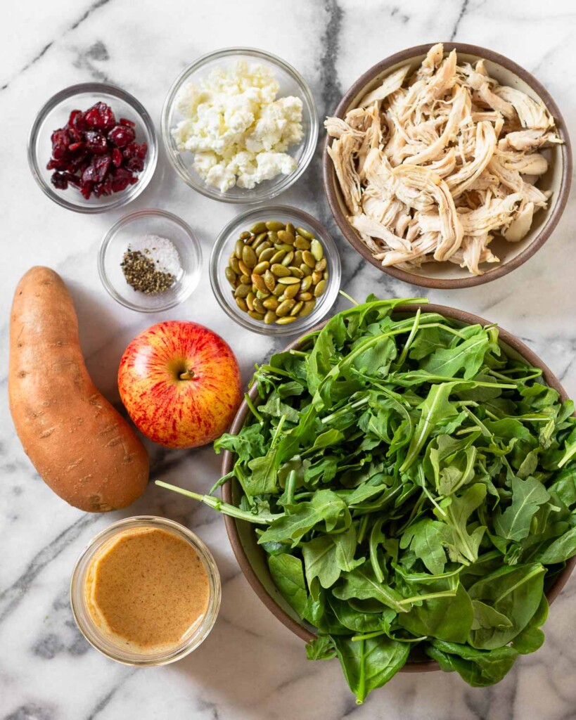 A bowl of shredded chicken, a bowl of mixed greens, a jar of dressing, a whole sweet potato, a whole red apple, a bowl of pumpkin seeds, a bowl of salt and pepper, a bowl of dried cranberries, and a bowl of goat cheese on a white marble countertop.