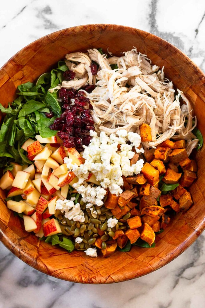 Large wooden salad bowl with shredded chicken, roasted diced sweet potatoes, pepitas, diced apples, dried cranberries, and goat cheese over mixed greens.