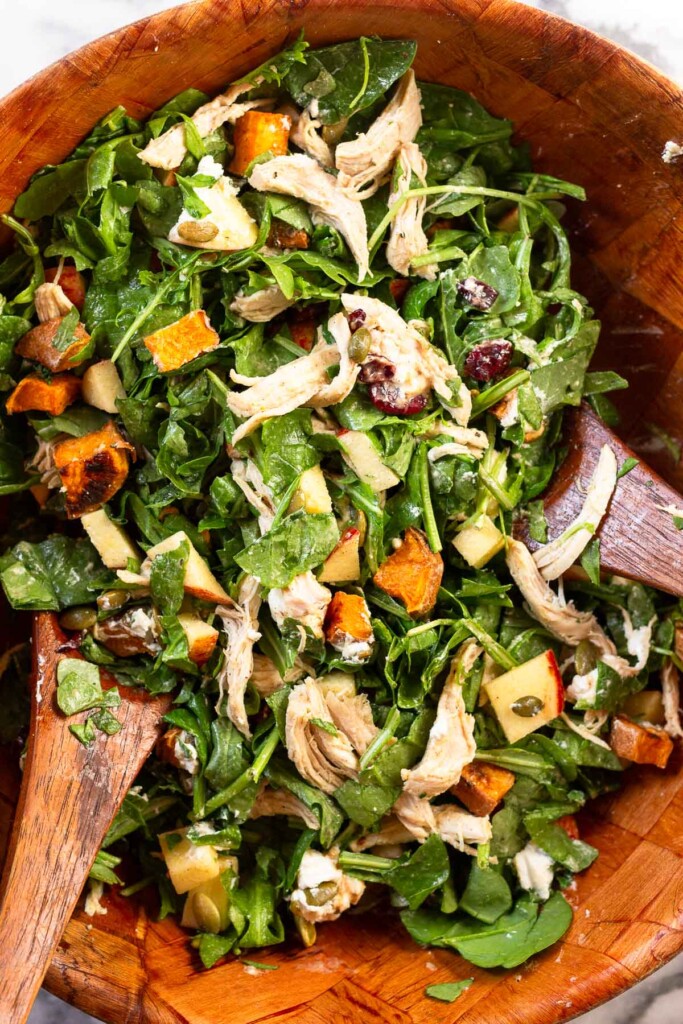 Overhead shot of a large wooden bowl with a harvest chicken salad with mixed greens, sweet potatoes, and apples in it. There are two wooden salad spoons in the bowl as well.