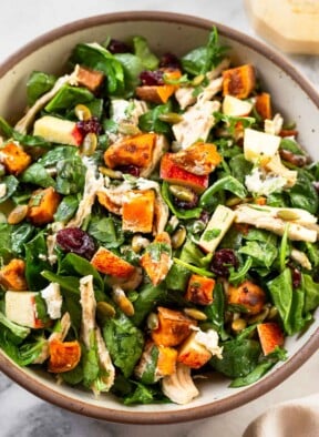 Chopped harvest salad with chicken, mixed greens, roasted sweet potatoes, dried cranberries, pumpkin seeds, and goat cheese all tossed with dressing in a white bowl. Next to the bowl is a jar of more dressing and a white kitchen towel.