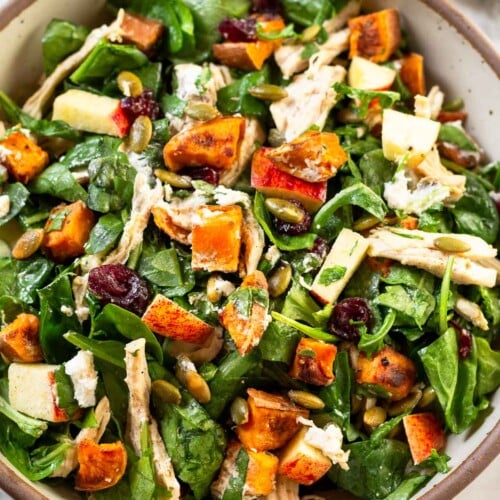 Chopped harvest salad with chicken, mixed greens, roasted sweet potatoes, dried cranberries, pumpkin seeds, and goat cheese all tossed with dressing in a white bowl. Next to the bowl is a jar of more dressing and a white kitchen towel.