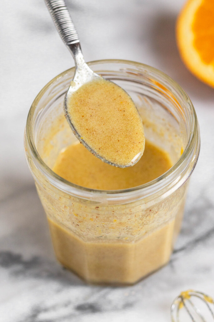 Orange vinaigrette in a jar with a spoon coming out of it. In front of the jar is a small whisk and behind the jar is half an orange.