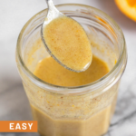 Orange vinaigrette in a jar with a spoon coming out of it. In front of the jar is a small whisk and behind the jar is half an orange.