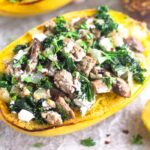 A turkey kale spaghetti squash boat sitting on a baking sheet. It is surrounded by half a lemon, a jar of red pepper flakes, and another spaghetti squash boat behind it.