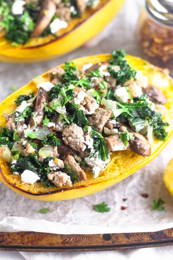A turkey kale spaghetti squash boat sitting on a baking sheet. It is surrounded by half a lemon, a jar of red pepper flakes, and another spaghetti squash boat behind it.