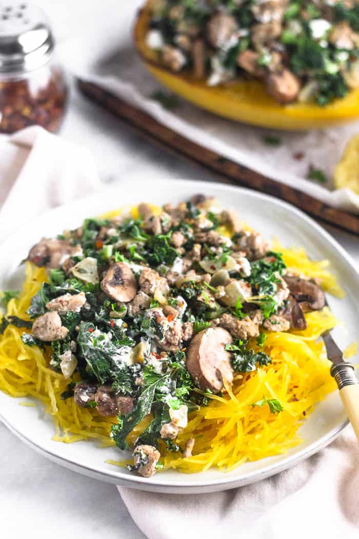 A white plate filled with spaghetti squash with ground turkey. A fork is resting on the plate and behind the plate is a baking sheet with turkey kale spaghetti squash boats and a jar of red pepper flakes.